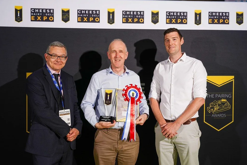 Three men holding a trophy and rosette at the Cheese Dairy Deli Expo awards backdrop