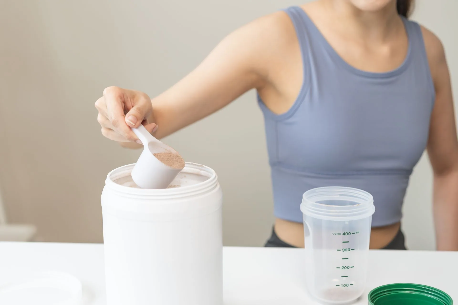 Woman scooping chocolate protein powder from a tub beside a measuring shaker
