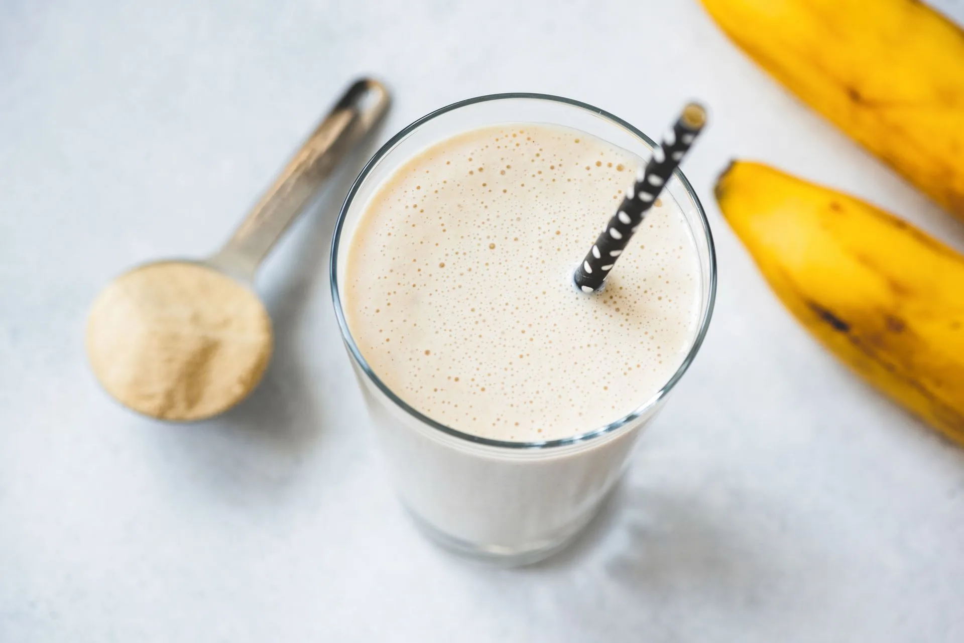 Glass of protein shake with a scoop of powder and two bananas on a pale surface