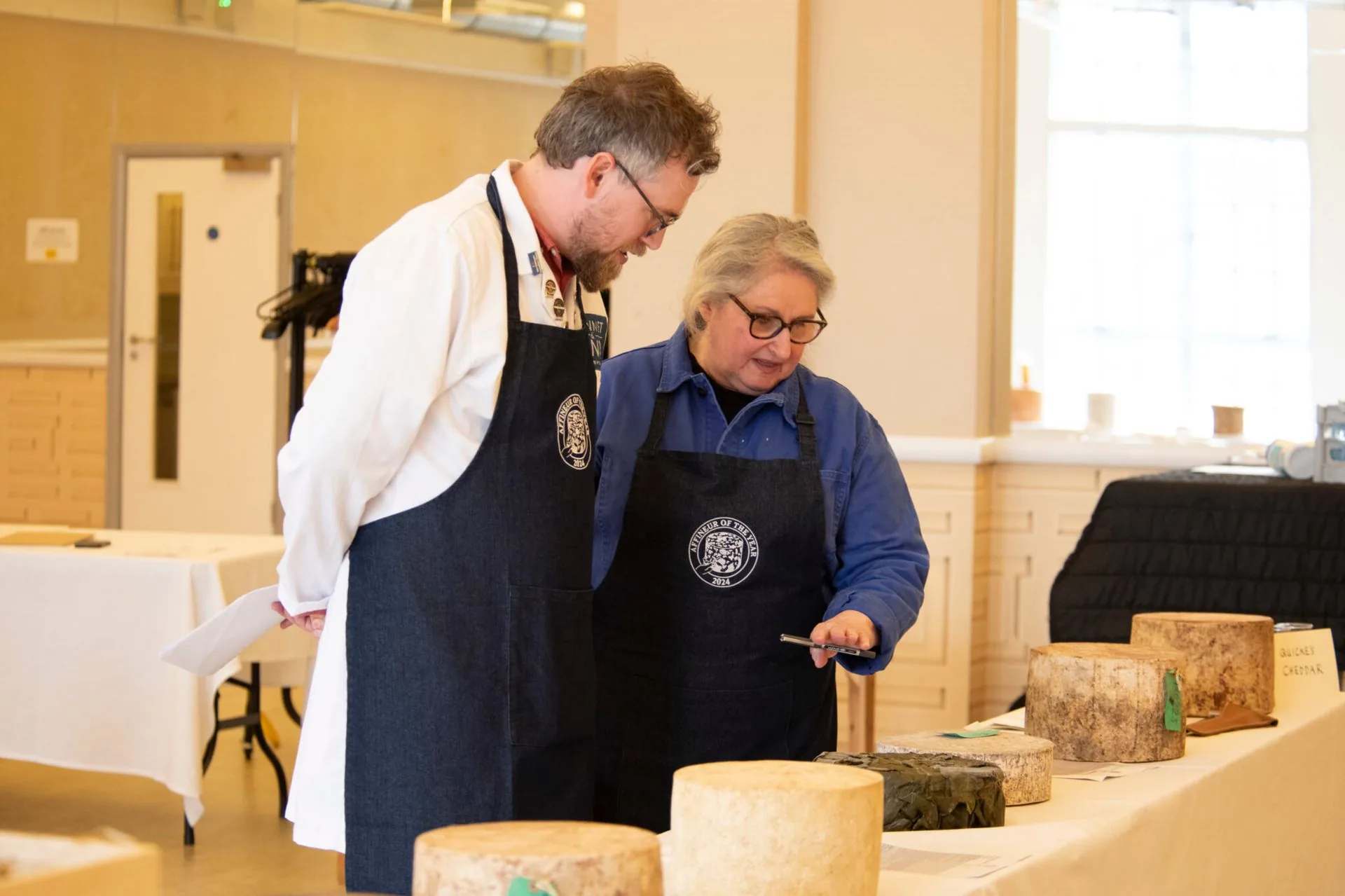 Two judges in Affineur of the Year aprons inspecting truckles of cheese on a table