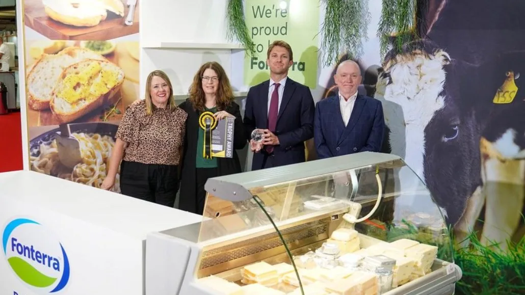 Four people at a Fonterra exhibition stand holding a glass trophy beside a cheese counter