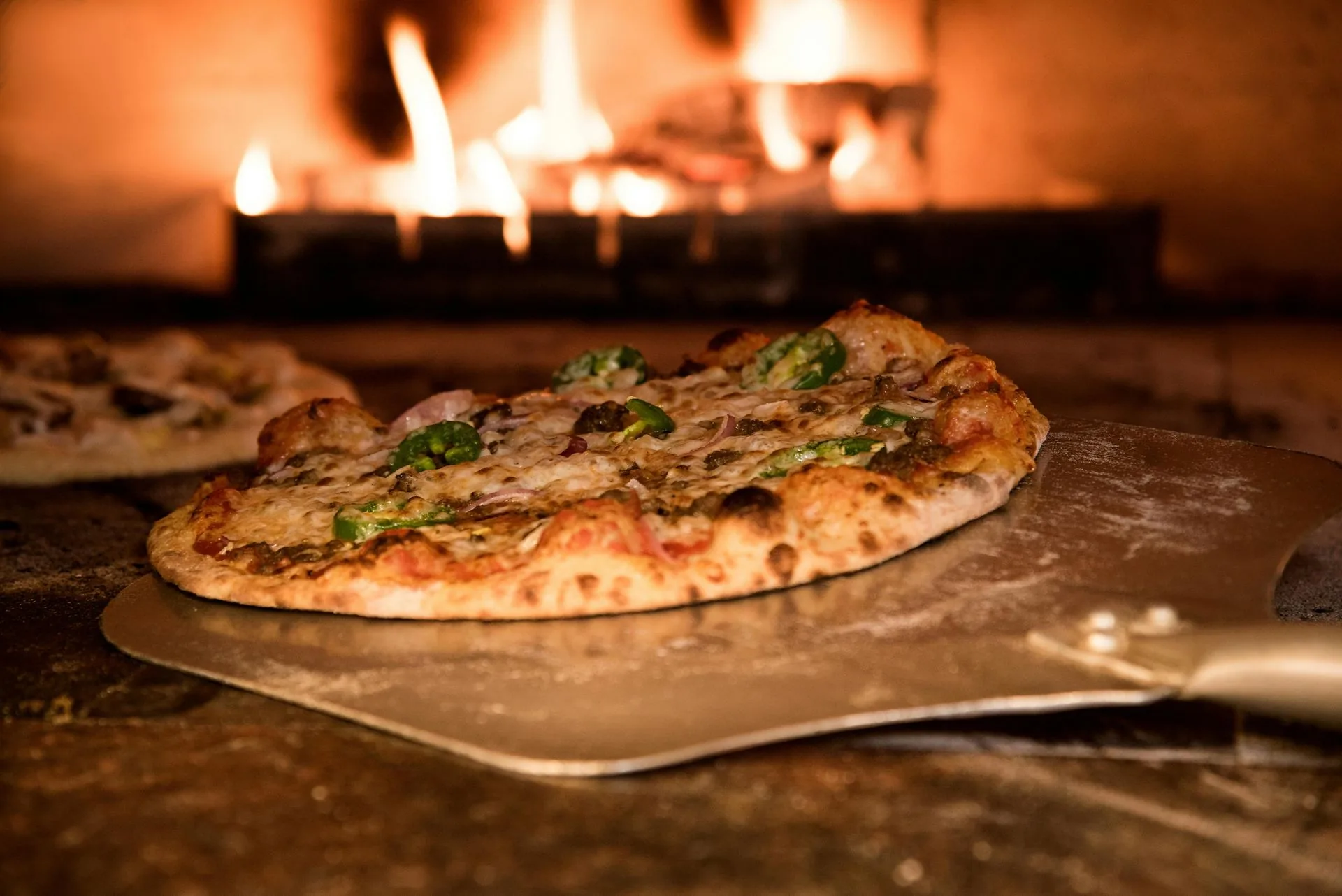 Wood-fired pizza with peppers on a peel before a flaming oven