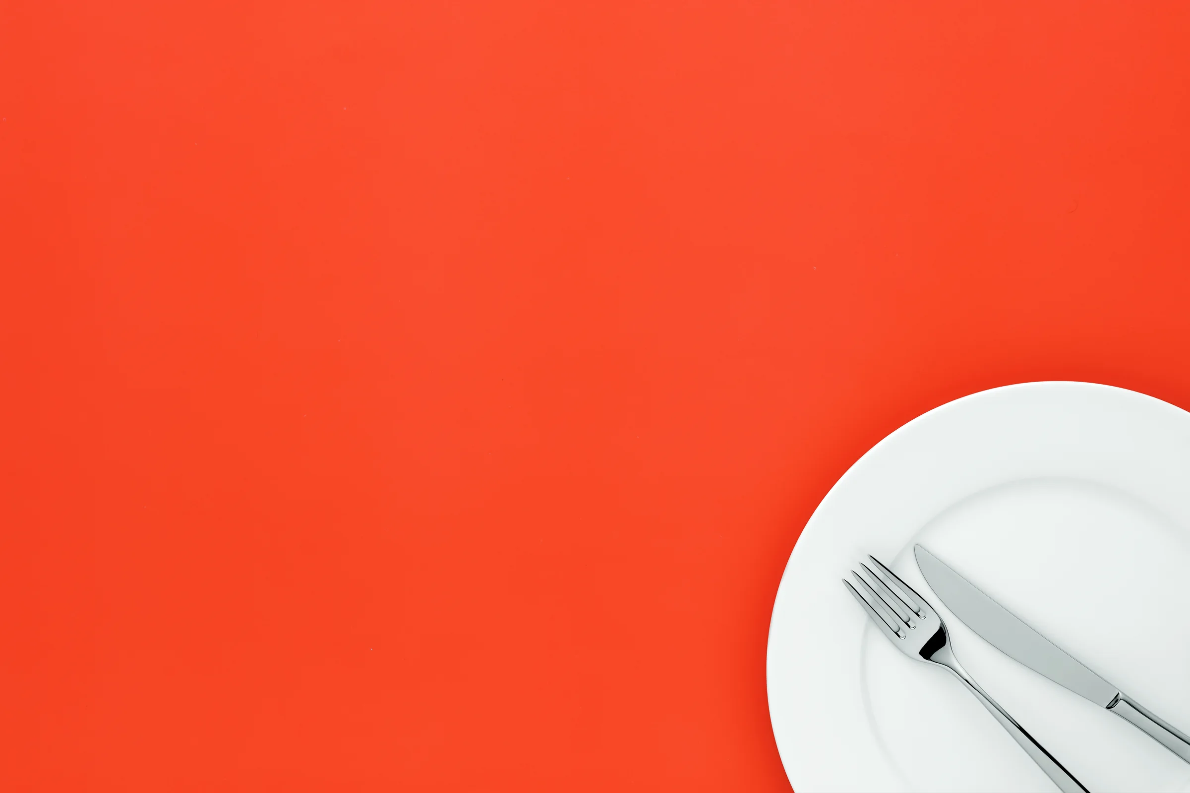 An empty white plate with a knife and fork on a bright orange table