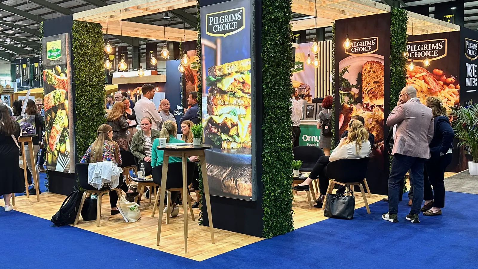 The Ornua stand: Pilgrims' Choice and Kerrygold lightboxes on foliage-clad columns, timber pergola and seating, with visitors sitting and talking